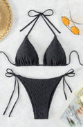 A two-piece black bikini set with a textured, dotted fabric pattern, featuring a halter triangle top and matching side-tie bottoms, laid flat on a white marble background with lifestyle props.