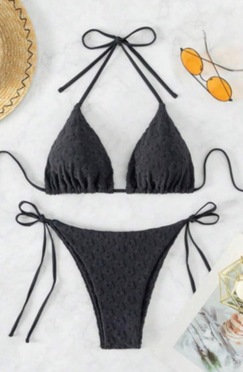 A two-piece black bikini set with a textured, dotted fabric pattern, featuring a halter triangle top and matching side-tie bottoms, laid flat on a white marble background with lifestyle props.